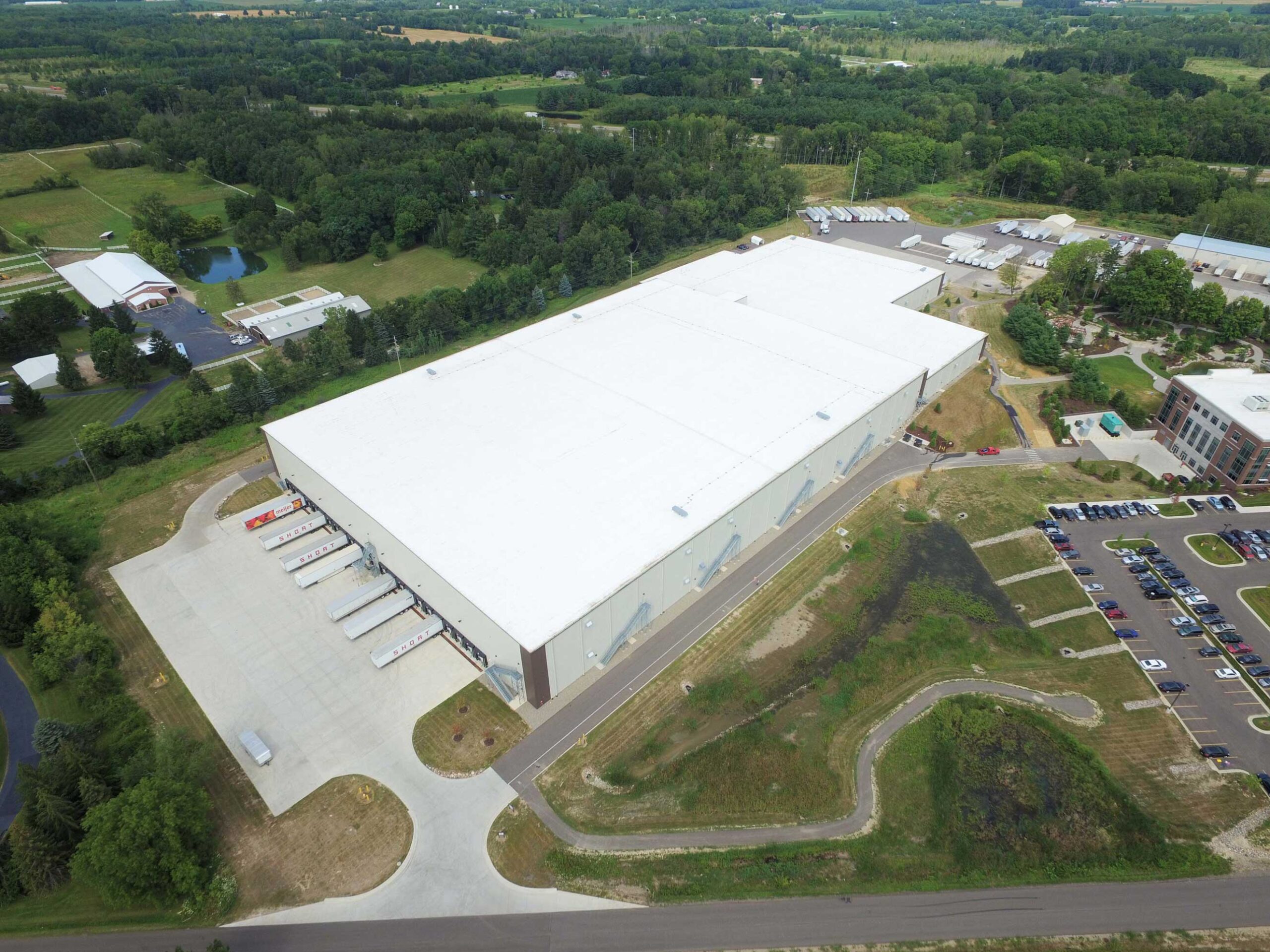 Aerial view of large warehouse with trucks parked alongside of it. Warehouse is surrounded by trees, parking lots, and other buildings.