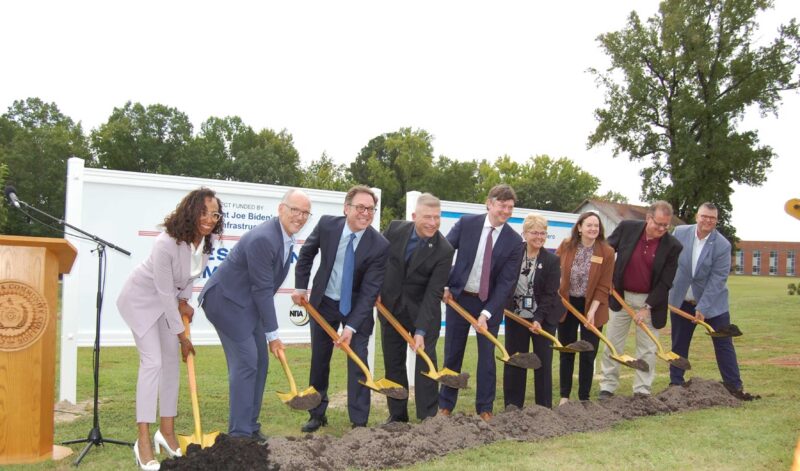 Eight people posing for a photo while scooping up dirt with their shovels.