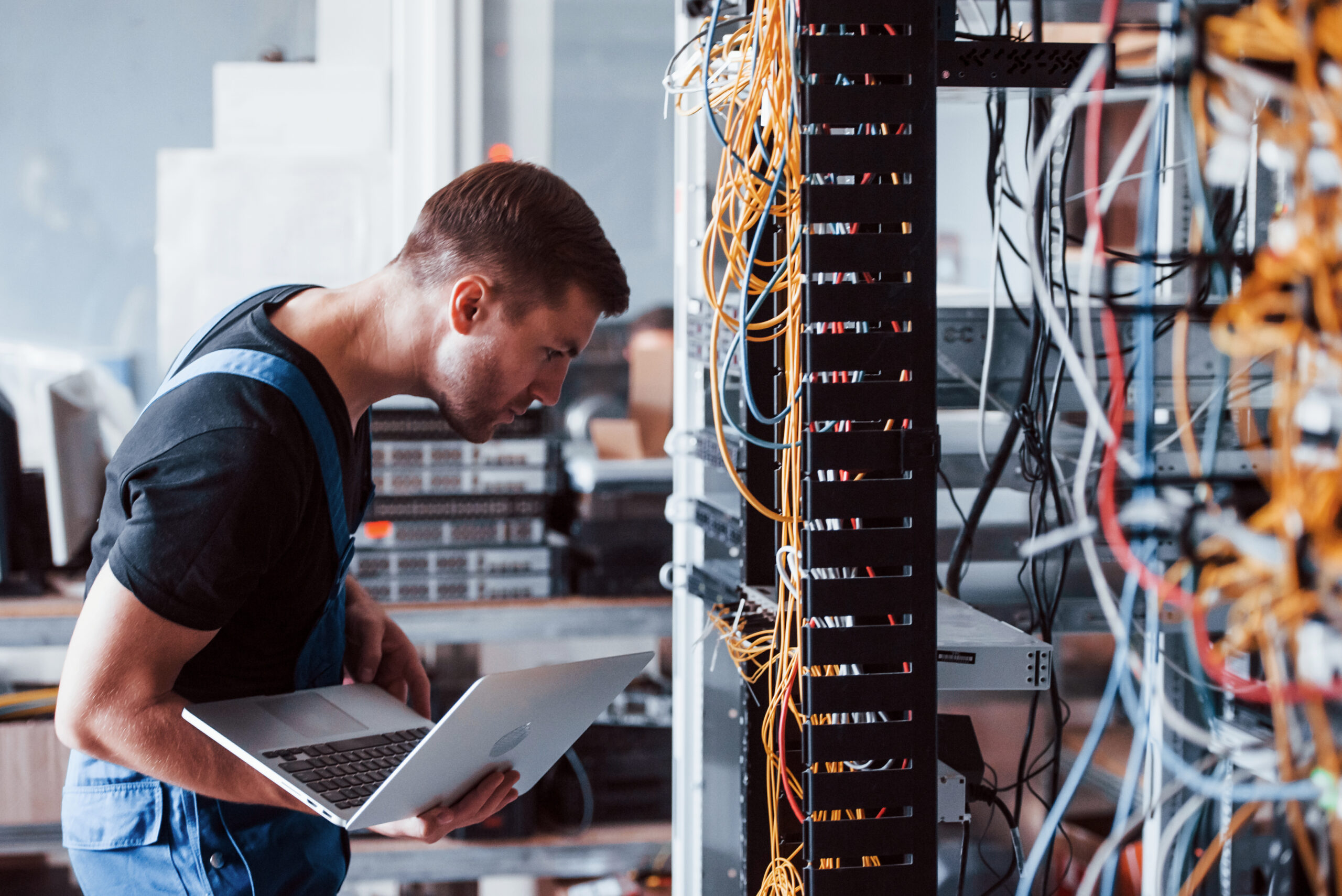 Young man holding a laptop evaluating various equipment and wires in a server room.