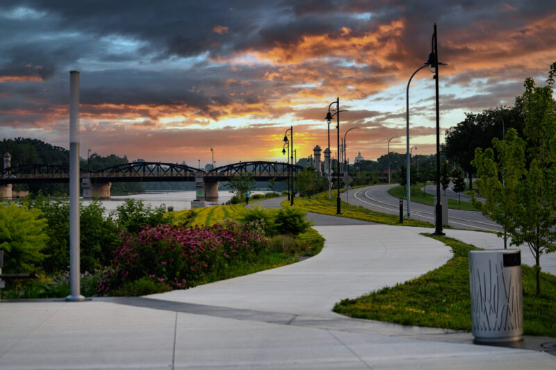 Winding sidewalk path at West River Wall in Rochester framed by flowers, greenery and new lighting and amenities.