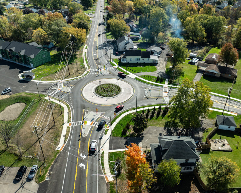 Vehicles driving around roundabout alongside homes and foliage.