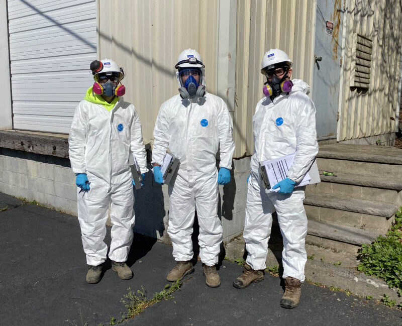 Three workers posing for a picture wearing white hazmat suits, latex gloves, and gas masks for protection.