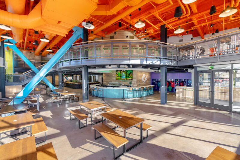 Interior of Mighty Squirrel Fenway showcases a bright orange ceiling and turquoise industrial beams. Picnic tables line the floor with a large center bar in the middle of the room.